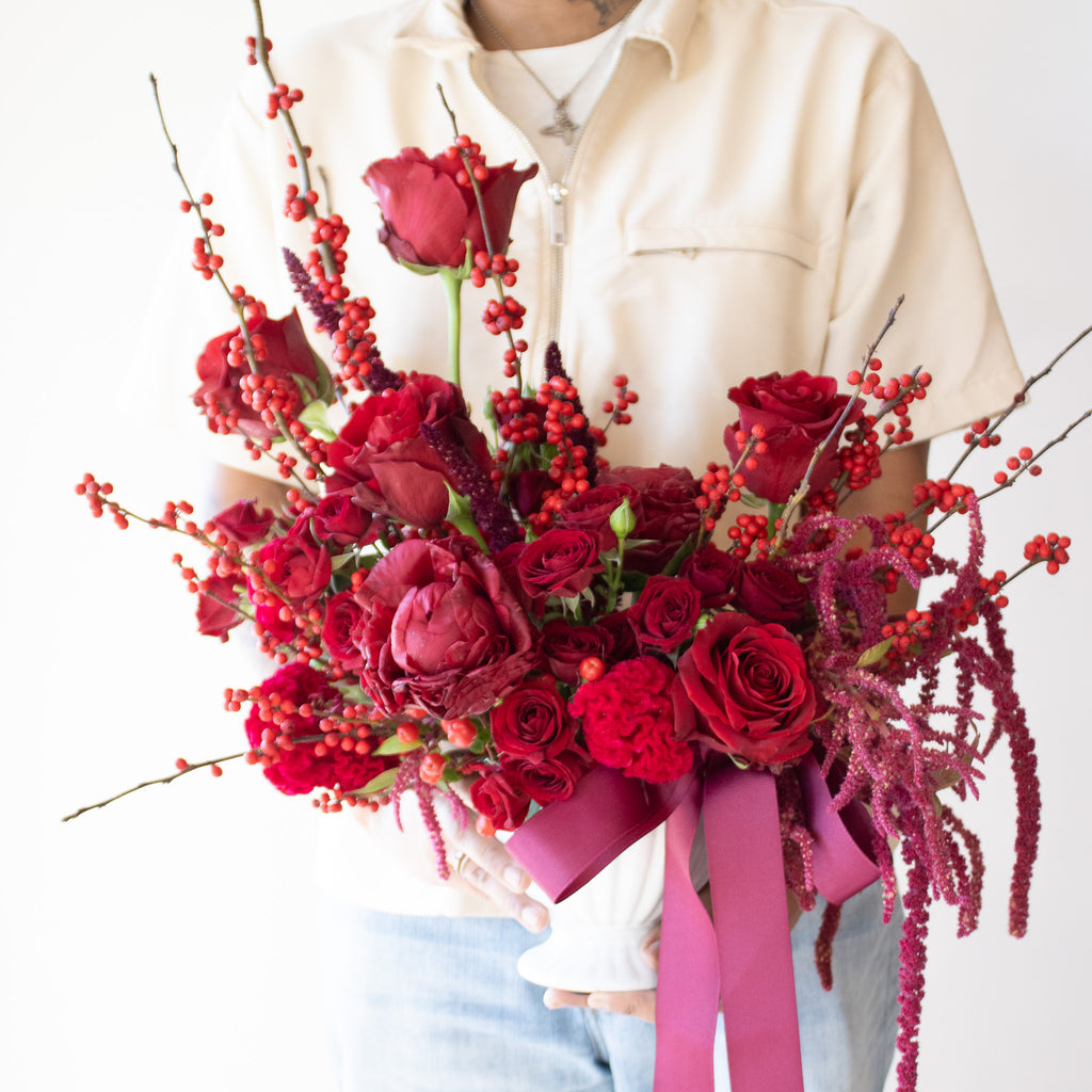 a person holding WildFlora's Red Velvet Arrangement in a white ceramic vase, wrapped with a fuchsia ribbon in front of a white backdrop. It has red flowers, including rose, spray rose, berries, amaranthus, peony, and celosia.
