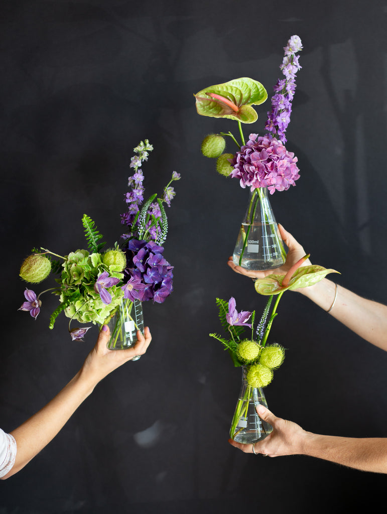 chartreuse and purple flowers in 3 glass beakers held by hands in front of a black background