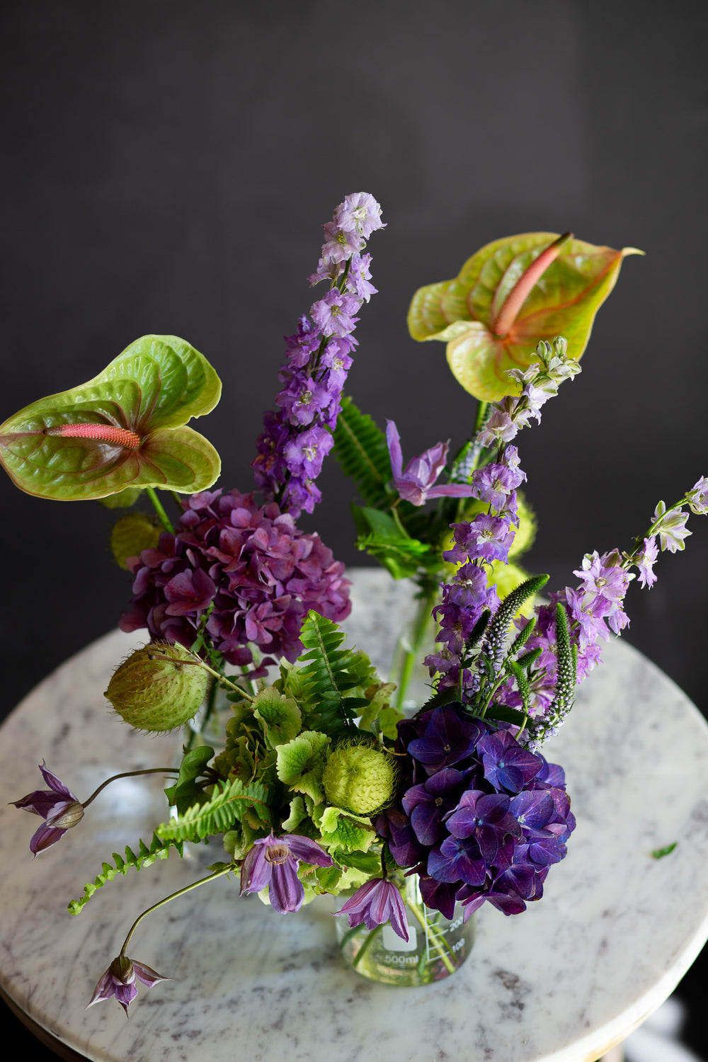 chartreuse and purple flowers in 3 glass beakers on a marble table against a dark background