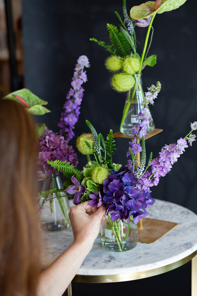 a woman adjusts chartreuse and purple flowers in 3 glass beakers on a marble table against a dark background