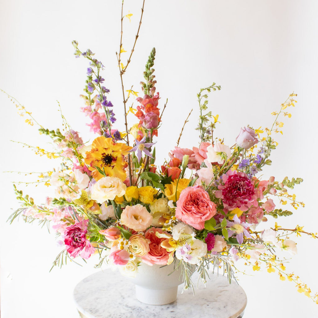 WildFlora's Stuck on Spring Arrangement in a white compote vase on a small marble table in front of a white backdrop. It has pink, peach, white, yellow, magenta, purple and white flowers, and includes rose, Japanese butterfly ranunculus, peony, delphinium, oncidium orchid, forsythia branch, lisianthus, clematis, sweet pea, pitcher flower, and snapdragon.