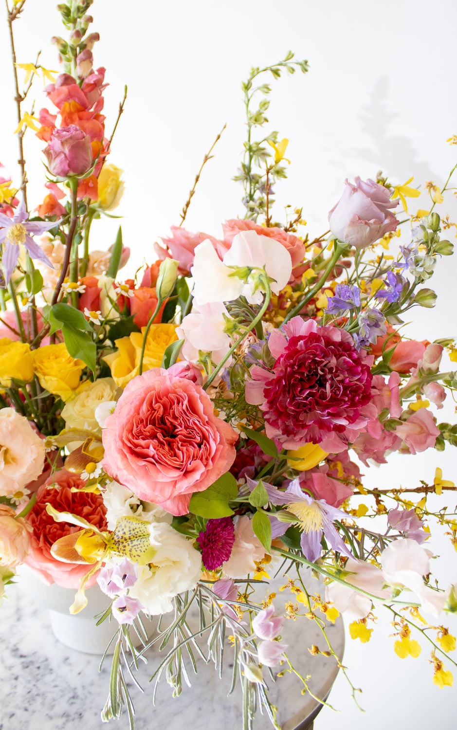 WildFlora's Stuck on Spring Arrangement in a white compote vase on a small marble table in front of a white backdrop. It has pink, peach, white, yellow, magenta, purple and white flowers, and includes rose, Japanese butterfly ranunculus, peony, delphinium, oncidium orchid, forsythia branch, lisianthus, clematis, sweet pea, pitcher flower, and snapdragon.