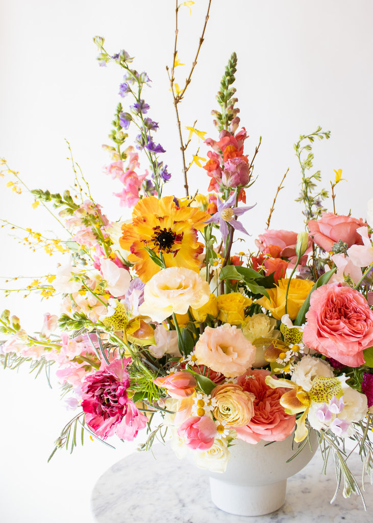 WildFlora's Stuck on Spring Arrangement in a white compote vase on a small marble table in front of a white backdrop. It has pink, peach, white, yellow, magenta, purple and white flowers, and includes rose, Japanese butterfly ranunculus, peony, delphinium, oncidium orchid, forsythia branch, lisianthus, clematis, sweet pea, pitcher flower, and snapdragon.