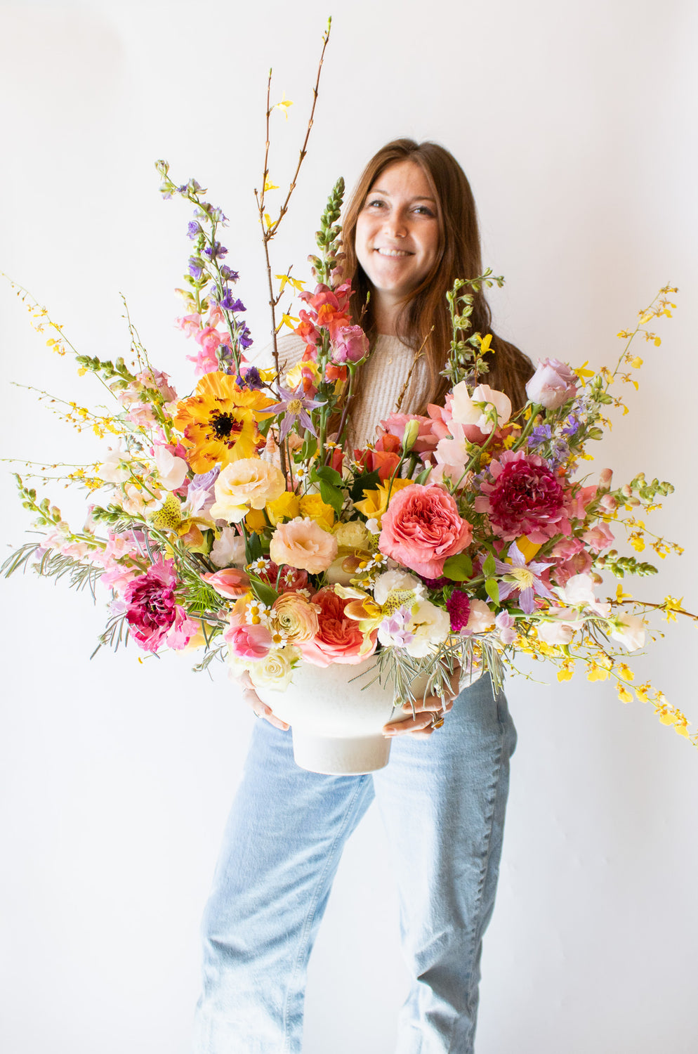 Woman holding a WildFlora's Stuck on Spring Arrangement in a white compote vase in front of a white backdrop. It has pink, peach, white, yellow, magenta, purple and white flowers, and includes rose, Japanese butterfly ranunculus, peony, delphinium, oncidium orchid, forsythia branch, lisianthus, clematis, sweet pea, pitcher flower, and snapdragon.