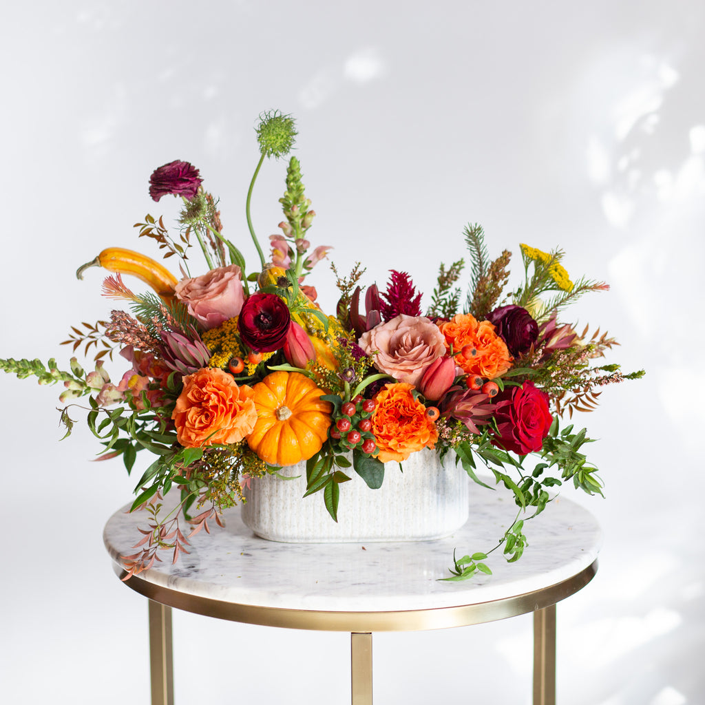 WildFlora's Gathered Garden Arrangement in a ribbed long and low white speckled container on a marble table. It includes orange, tangerine, red, pink, fuchsia, maroon, and gold flowers, complete with festive gourds / mini pumpkin, ranunculus, rose, dahlia, yarrow, weeping amaranthus, chocolate cosmos, delphinium, lisianthus, tulip, Queen Anne's lace, evergreens, snapdragon, and hypericum berries.