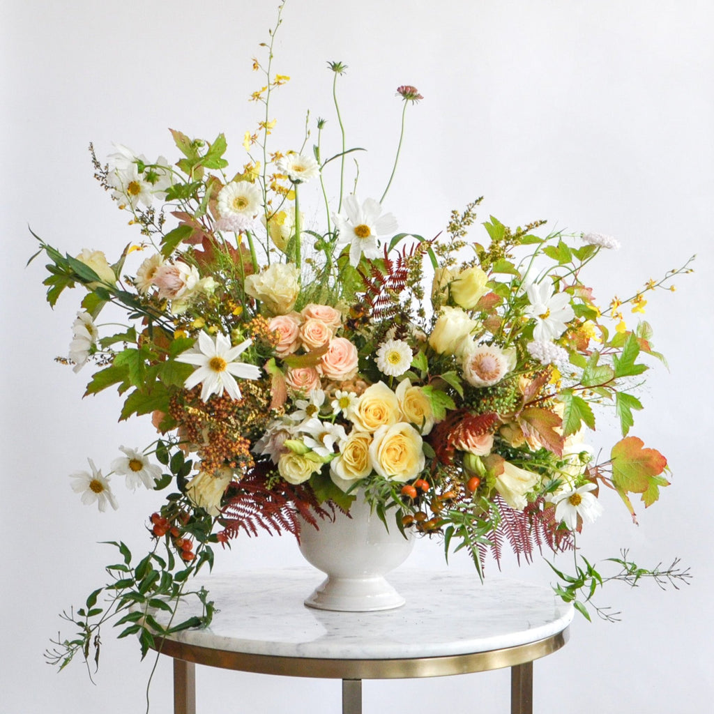 A green, yellow, peach, and maroon WildFlora flower arrangement in a white vase, featuring rose, lisianthus, fern, and oncidium orchid, cosmos, fall foliage leaves, protea, gerber daisy, and berries.