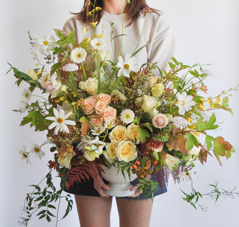 A woman holding a green, yellow, peach, and maroon WildFlora flower arrangement in a white vase, featuring rose, lisianthus, fern, and oncidium orchid, cosmos, fall foliage leaves, blushing bride protea, allium, gerber daisy, and berries.