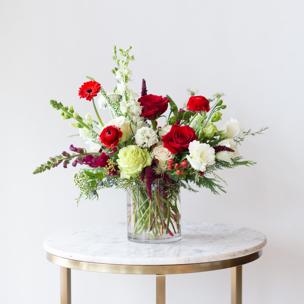 WildFlora's Holiday Cheer Arrangement in a glass vase on a small marble table in front of a white backdrop. It has red, green, white, and chartreuse flowers, and includes rose, delphinium, snapdragon, scabiosa, piccolini daisy, berries, and lisianthus.