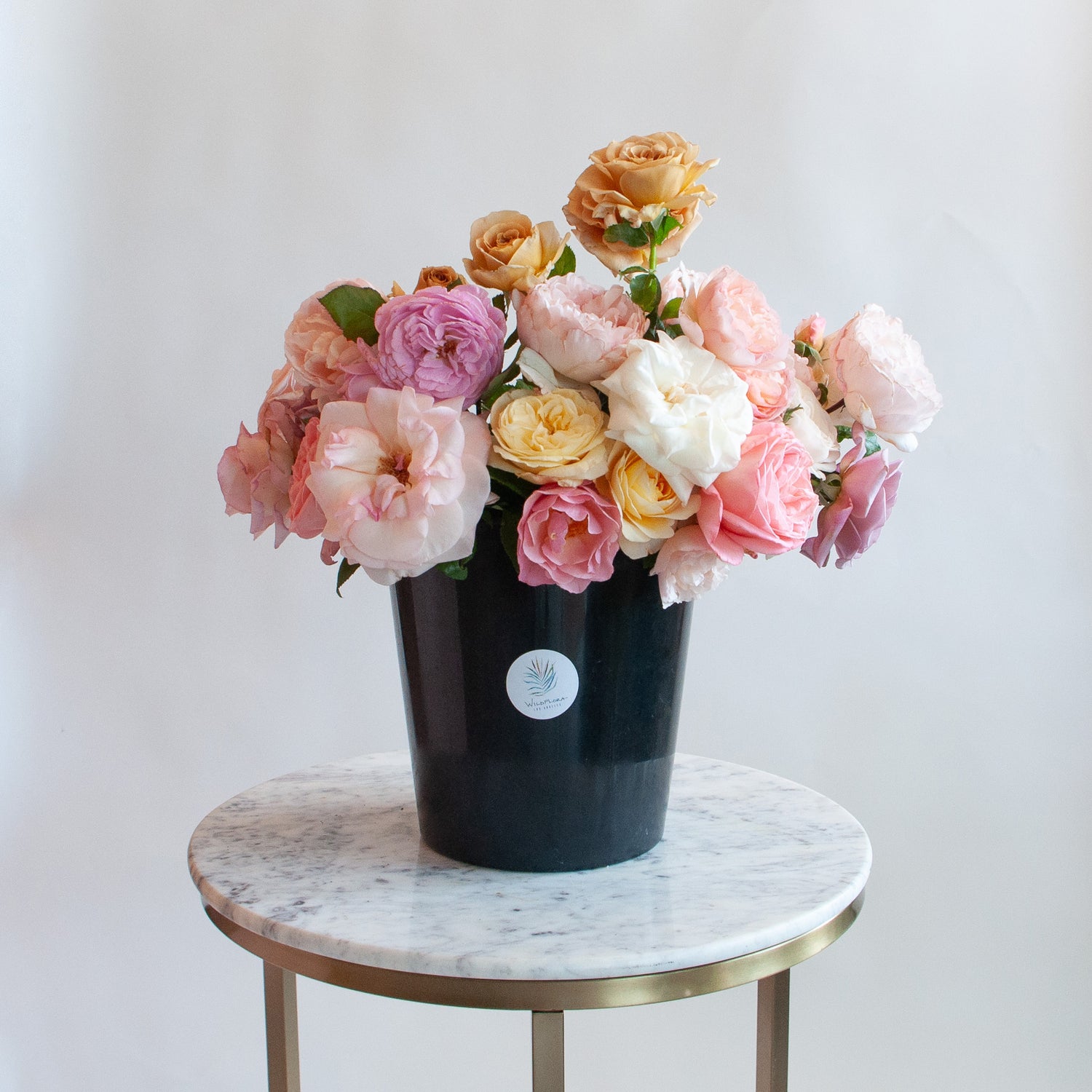 A bunch of organic roses in a black bucket in front of a white backdrop on a marble table. It's filled with pink, yellow, lavender, and and peach roses, including multiple varieties.