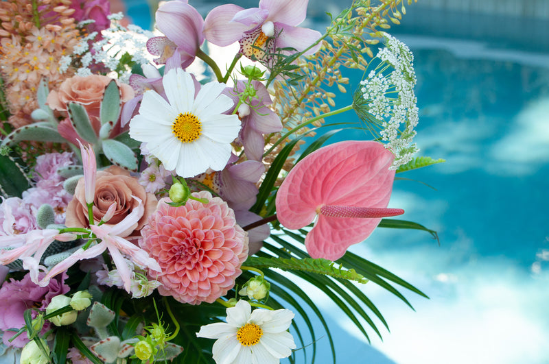 Closeup of a very large pink, peach, white, and green flower arrangement. The design is inspired by both the desert and lush vibes of Palm Springs, California, and includes cacti, succulents, roses, anthurium, cosmos, lisianthus, Queen Anne's lace, foxtail lily, orchids, snapdragon, palm fronds, and monstera leaves in a round terracotta pot.