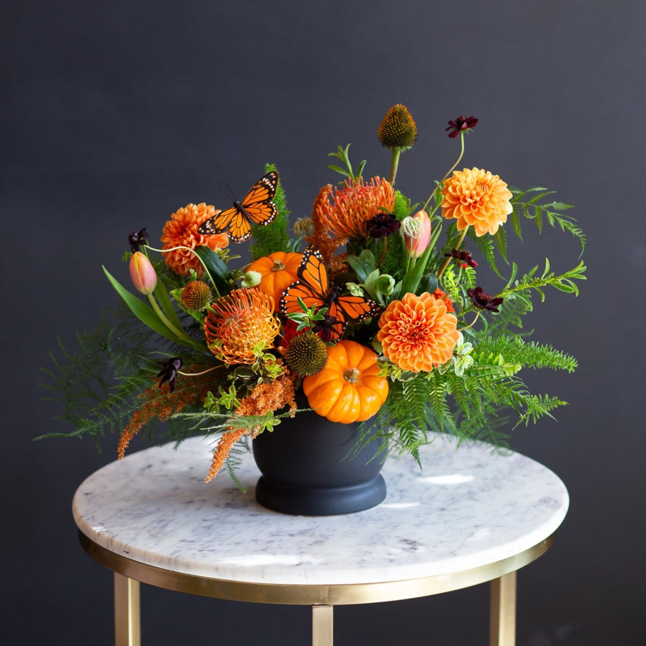 orange dahlias, pincushion protea, and other orange flowers in a sprawling bed of greens in a black container on a marble table in front of a black backdrop. There are also tiny orange pumpkins and monarch butterfly decals in this WildFlora arrangement.