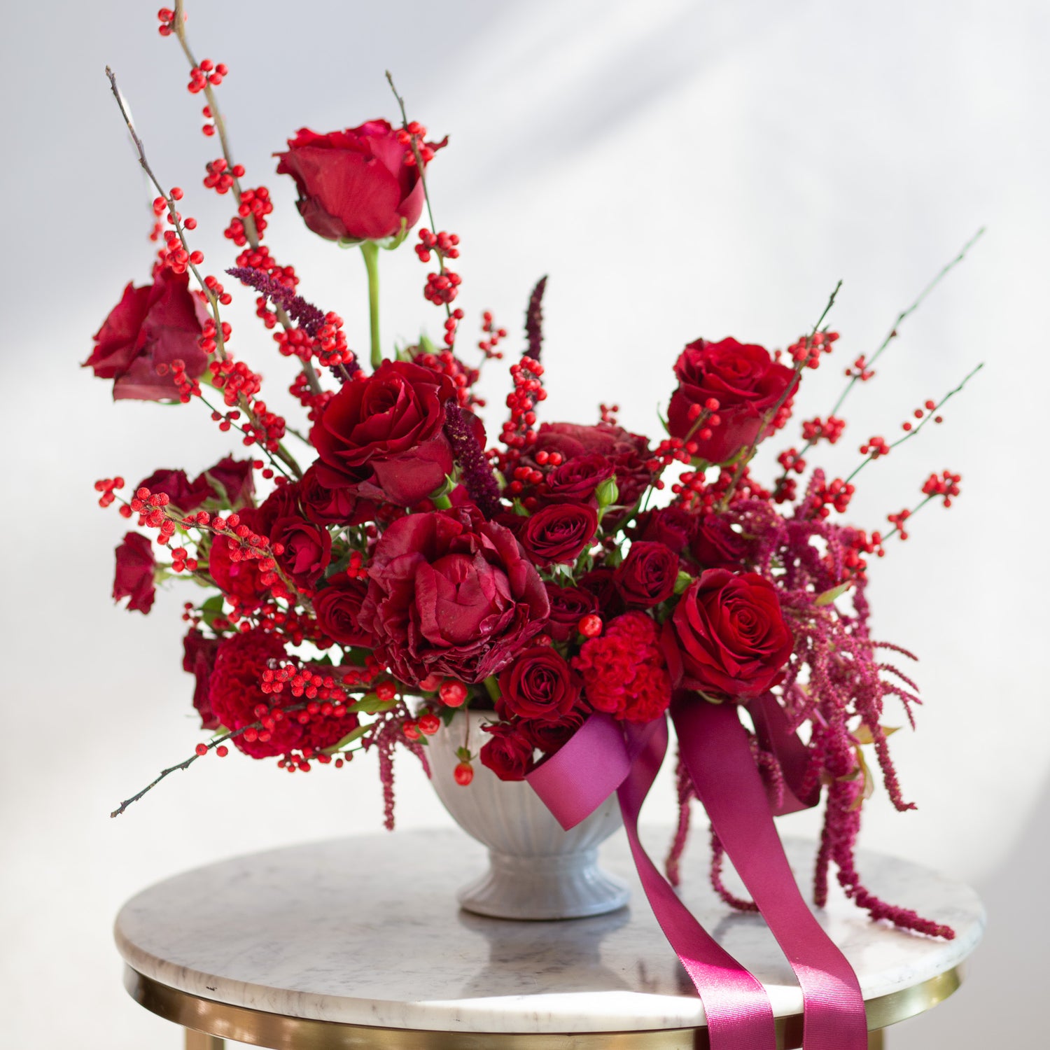 WildFlora's Red Velvet Arrangement in a white ceramic vase, wrapped with a fuchsia ribbon, on a small marble table in front of a white backdrop. It has red flowers, including rose, spray rose, berries, amaranthus, peony, and celosia.