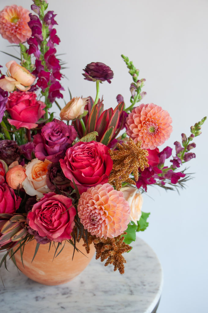 closeup of A red, peach, pink, and eggplant WildFlora flower arrangement in a spherical terracotta vase, featuring rose, ranunculus, dahlia, lisianthus, amaranthus, and snapdragon.