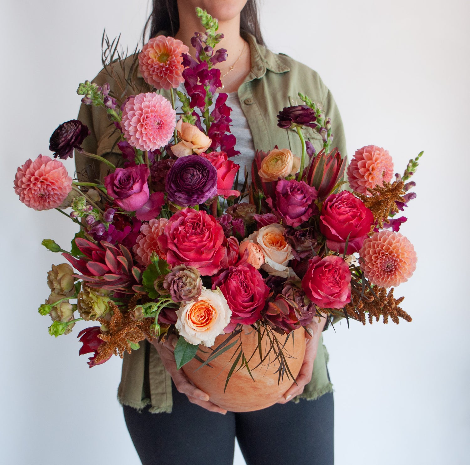 woman holding A red, peach, pink, and eggplant WildFlora flower arrangement in a spherical terracotta vase, featuring rose, ranunculus, dahlia, lisianthus, amaranthus, and snapdragon.