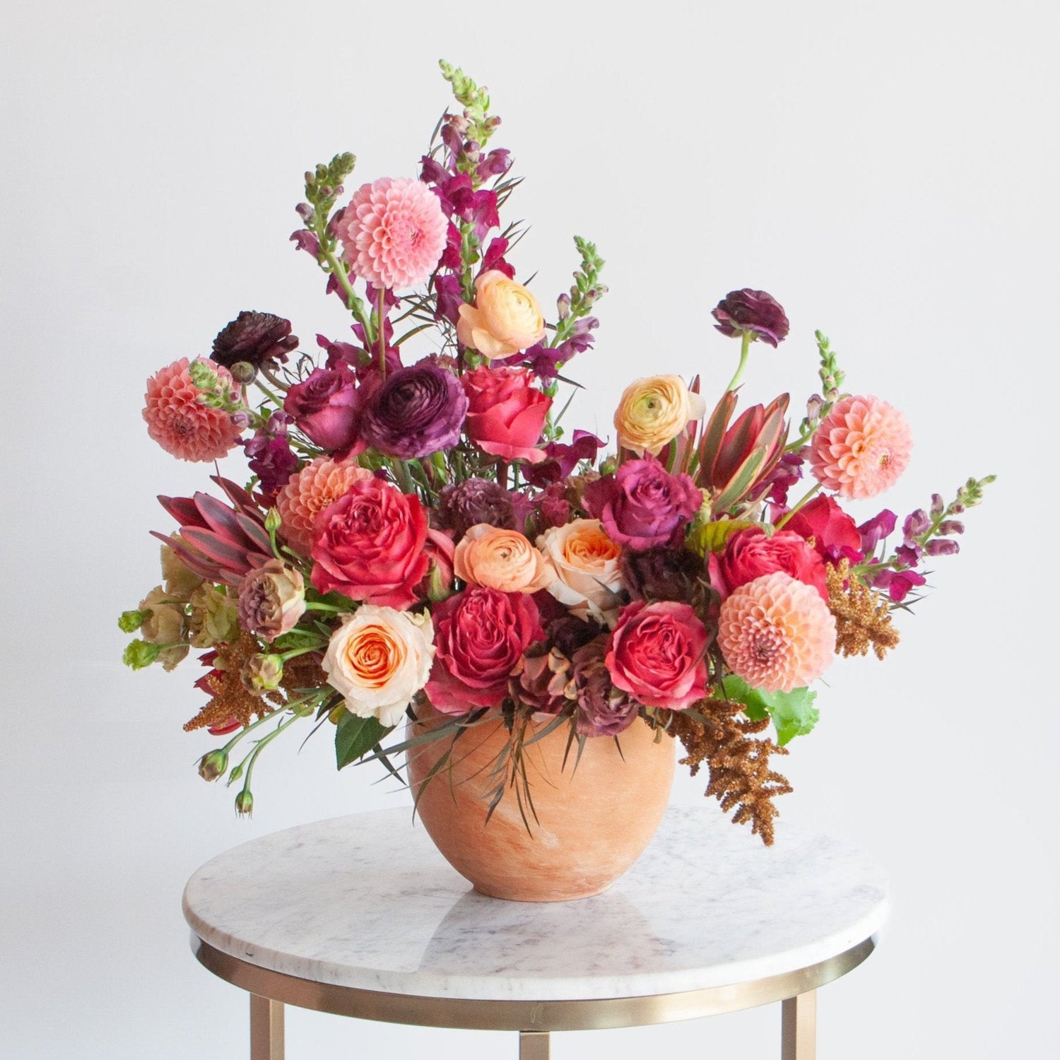 A red, peach, pink, and eggplant WildFlora flower arrangement in a spherical terracotta vase, featuring rose, ranunculus, dahlia, lisianthus, amaranthus, and snapdragon.