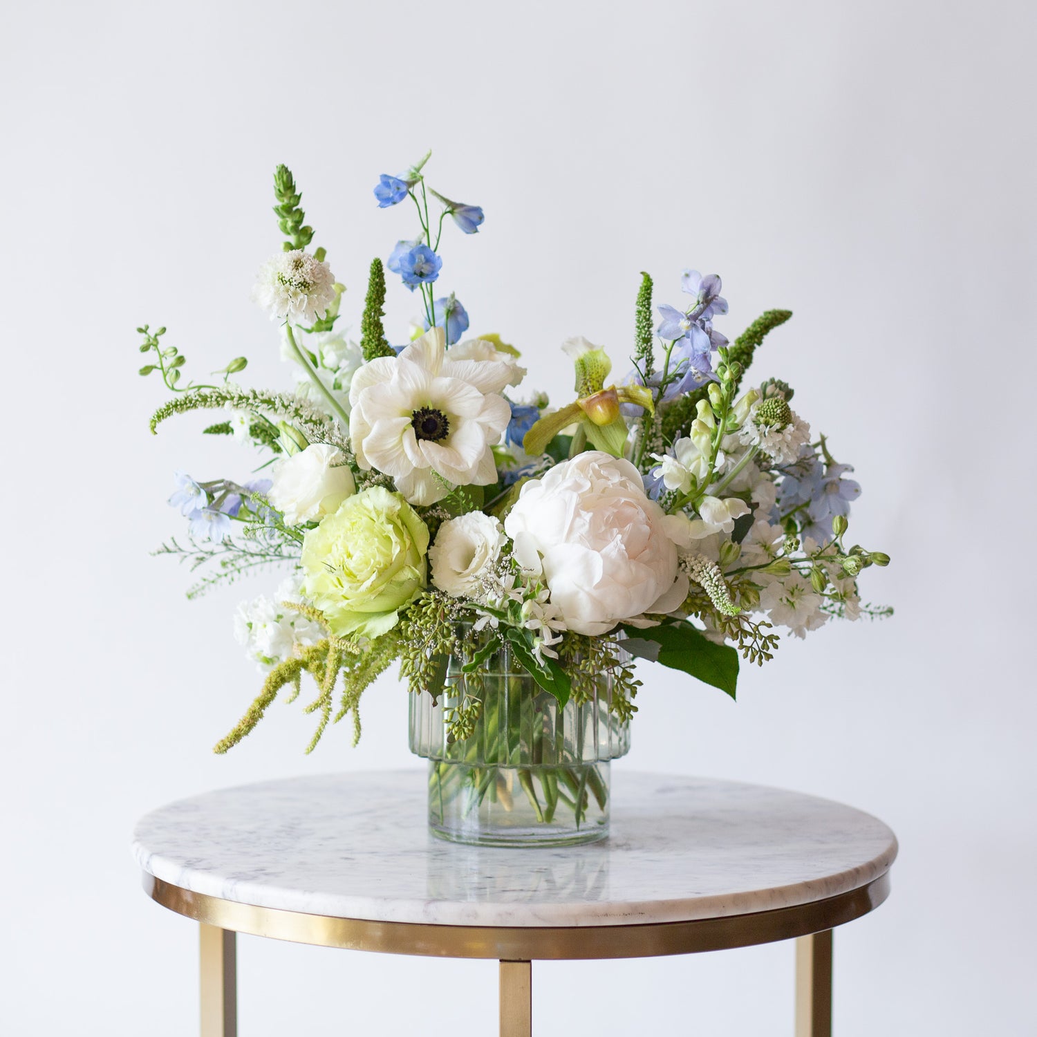 WildFlora's Starry Solstice Arrangement in a ribbed glass vase on a small marble table in front of a white backdrop. It has baby blue, green, and white flowers, and includes rose, veronica, lisianthus, lily, peony, snapdragon, delphinium, scabiosa, amaranthus, anemone, and pitcher flower. 