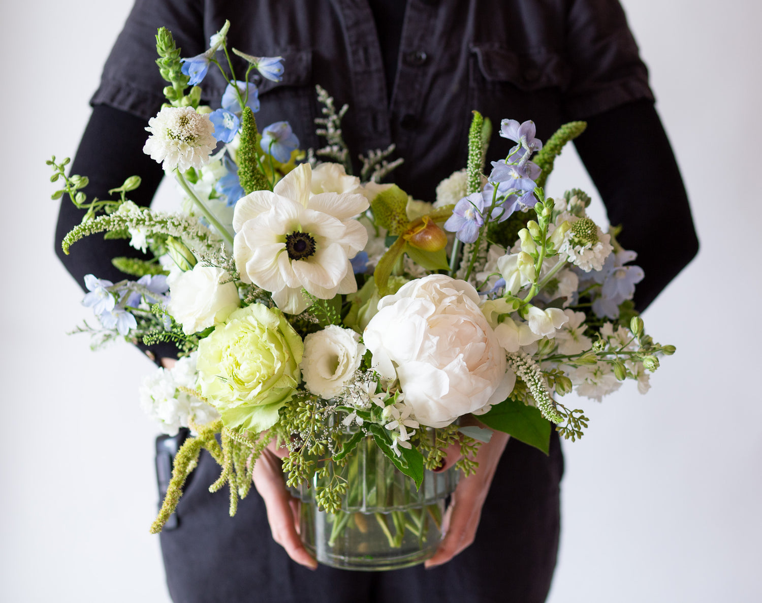 woman holding WildFlora's Starry Solstice Arrangement in a ribbed glass vase in front of a white backdrop. It has baby blue, green, and white flowers, and includes rose, veronica, lisianthus, lily, peony, snapdragon, delphinium, scabiosa, amaranthus, anemone, and pitcher flower. 