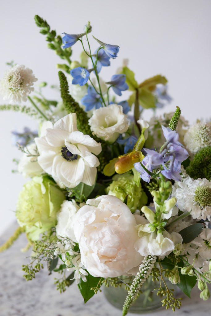 closeup of WildFlora's Starry Solstice Arrangement in a ribbed glass vase on a small marble table in front of a white backdrop. It has baby blue, green, and white flowers, and includes rose, veronica, lisianthus, lily, peony, snapdragon, delphinium, scabiosa, amaranthus, anemone, and pitcher flower. 