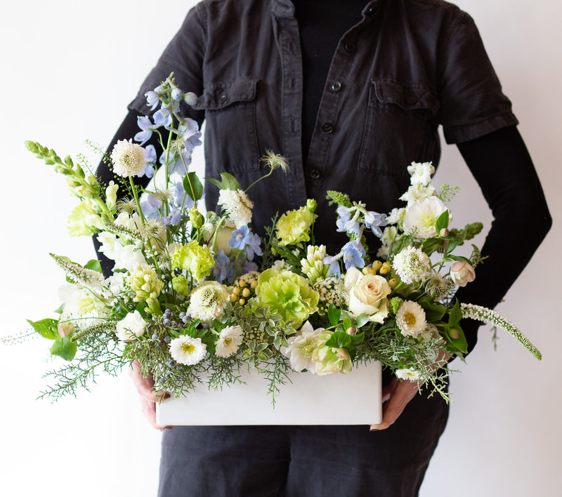 Woman holding a WildFlora Flora Flurries Long & Low Arrangement in a white ceramic long & low vase in front of a white backdrop. It has baby blue, chartreuse, green, and white flowers, and includes rose, veronica, lisianthus, snapdragon, hyacinth, delphinium, scabiosa, juniper, aster, and hellebore.