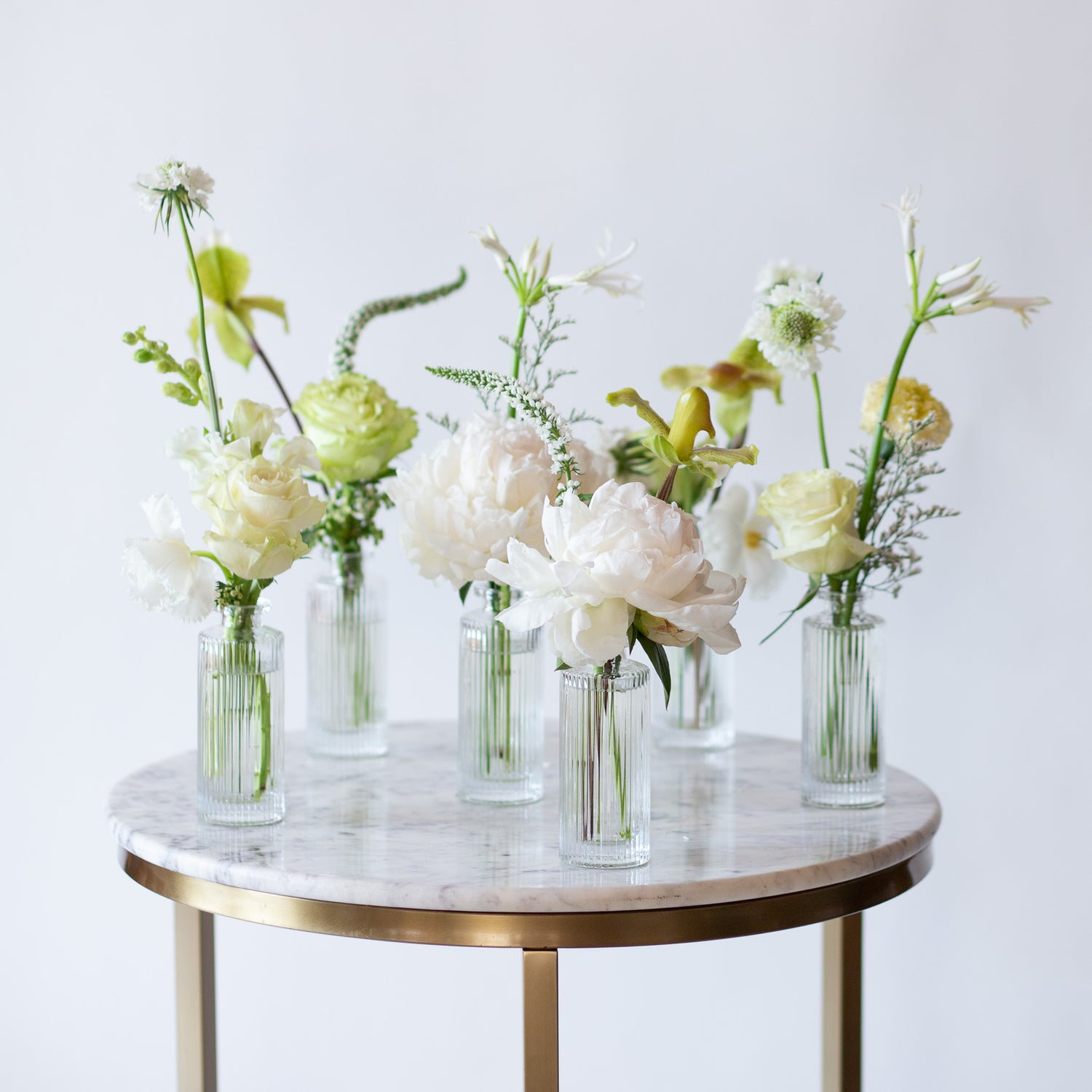 WildFlora's Snowy Days Bud Vases on a small marble table in front of a white backdrop. They have green, white, and chartreuse flowers, and include rose, veronica, scabiosa, pitcher flower, lily, and peony.