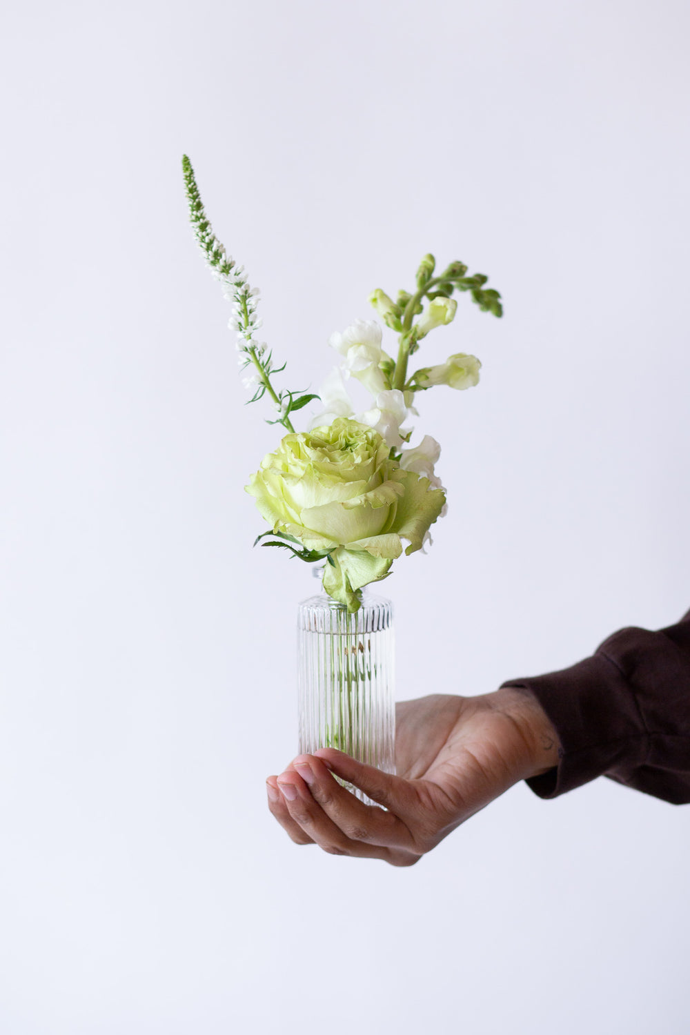 A hand holding one of WildFlora's Snowy Days Bud Vases in front of a white backdrop. They have green, white, and chartreuse flowers, and include rose, veronica, scabiosa, pitcher flower, lily, and peony.