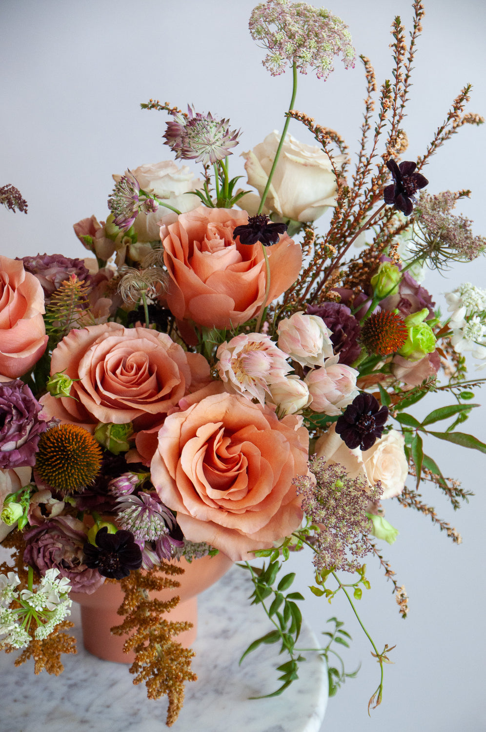 A cream, eggplant, salmon and peach WildFlora flower arrangement in a terracotta compote vase, featuring rose, thistle, Queen Anne's lace, blushing bride protea, and lisianthus.