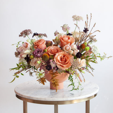 A cream, eggplant, salmon and peach WildFlora flower arrangement in a terracotta compote vase, featuring rose, thistle, Queen Anne's lace, blushing bride protea, and lisianthus.