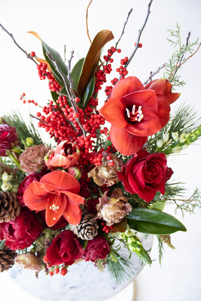 WildFlora's Winterberry Arrangement in a white ceramic compote vase on a small marble table in front of a white backdrop. It has red, green, white, and brown flowers, and includes rose, amaryllis, pinecones, peony, berries, lisianthus, snapdragon, and winter greens and magnolia foliage.