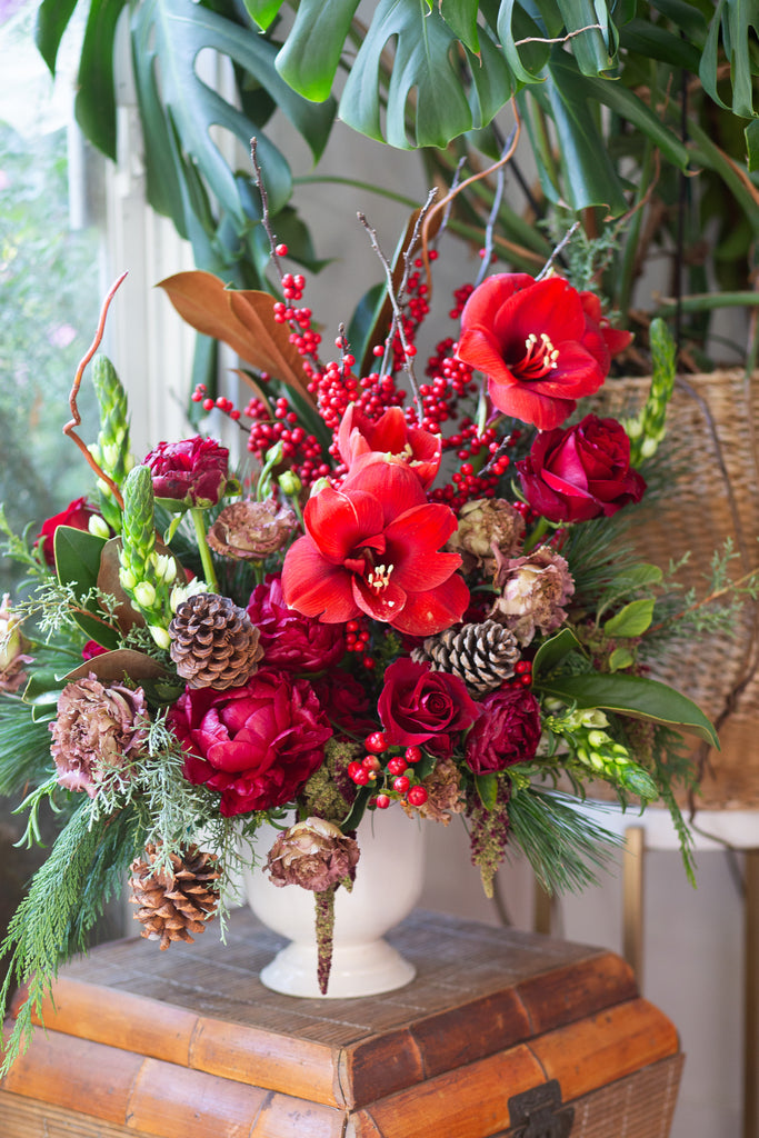 WildFlora's Winterberry Arrangement in a white ceramic compote vase on side table in a home. It has red, green, white, and brown flowers, and includes rose, amaryllis, pinecones, peony, berries, lisianthus, snapdragon, and winter greens and magnolia foliage.