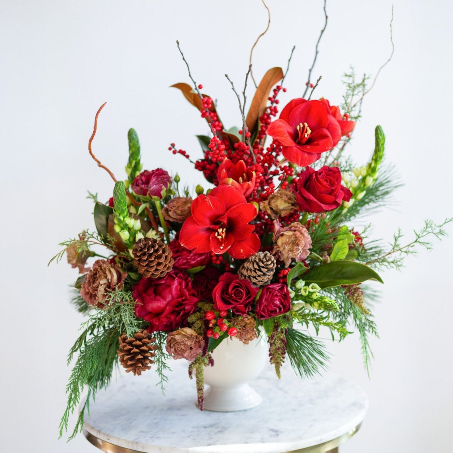 WildFlora's Winterberry Arrangement in a white ceramic compote vase on a small marble table in front of a white backdrop. It has red, green, white, and brown flowers, and includes rose, amaryllis, pinecones, peony, berries, lisianthus, snapdragon, and winter greens and magnolia foliage.