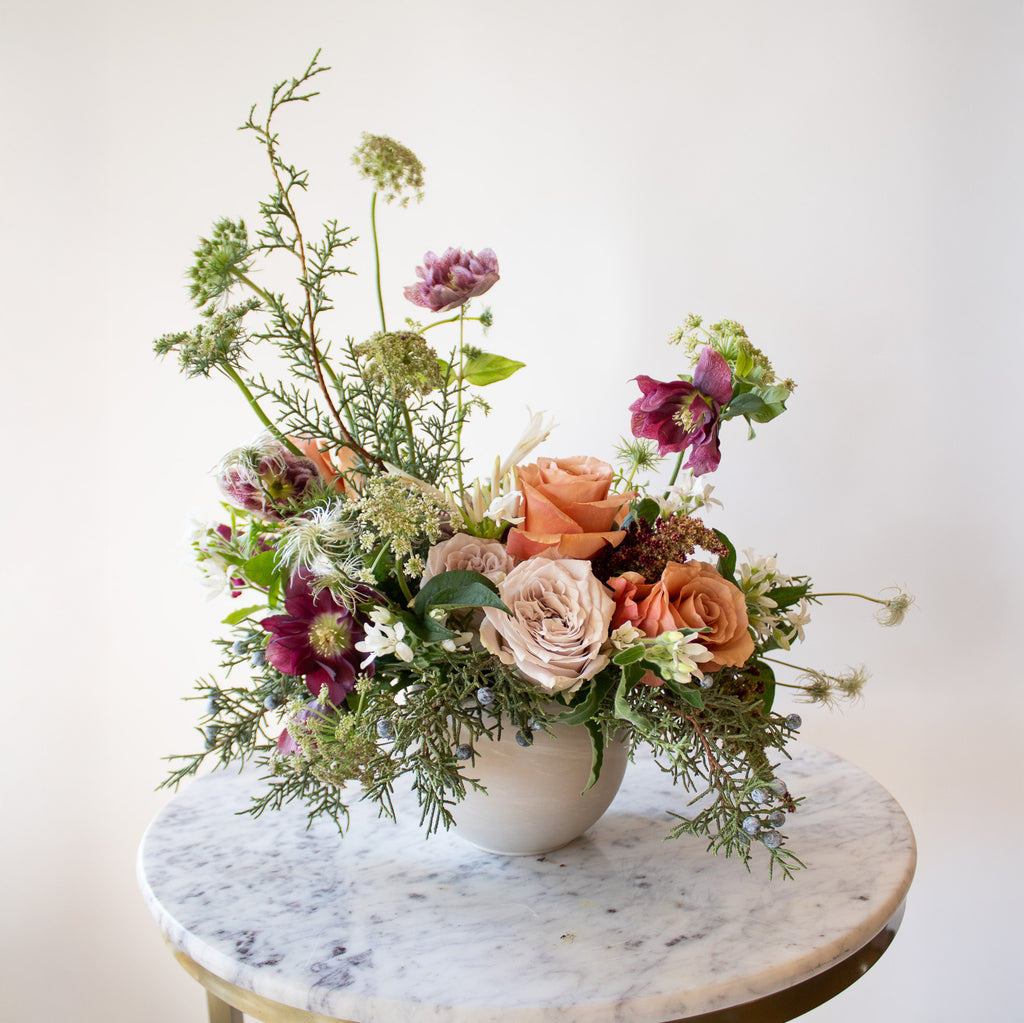 WildFlora's Winter's Garden Arrangement in a spherical grey vase, on a marble table in front of a white backdrop. It has sienna orange, pastel violet, red, pink, fuchsia, eggplant, and white flowers, and includes rose, scabiosa, hellebore, Queen Anne's lace, and juniper.