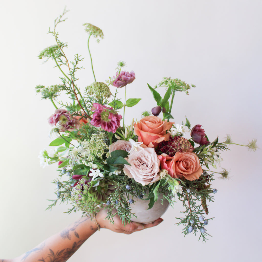 WildFlora's Winter's Garden Arrangement in a spherical grey vase, held by a hand in front of a white backdrop. It has sienna orange, pastel violet, red, pink, fuchsia, eggplant, and white flowers, and includes rose, scabiosa, hellebore, Queen Anne's lace, and juniper.