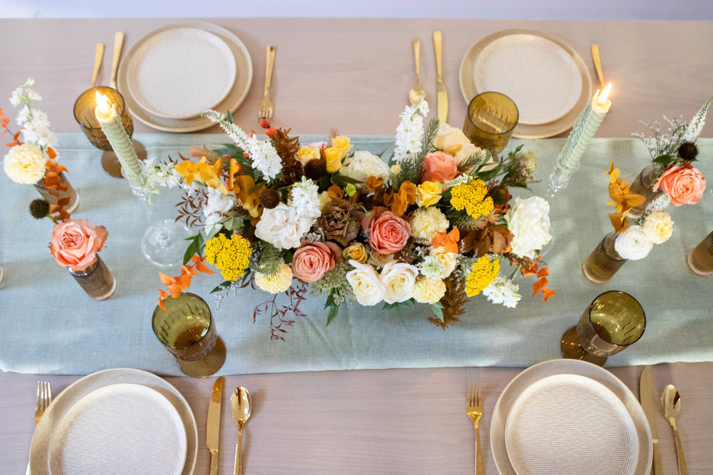 WildFlora's Gathered Garden Arrangement in an art deco white long & low container on a thanksgiving table. It include coral, yellow, gold, brown, orange, and white flowers and foliage, complete with roses, spray roses, weeping amaranthus, delphinium, lisianthus, echinacea, yarrow, and marigold. 