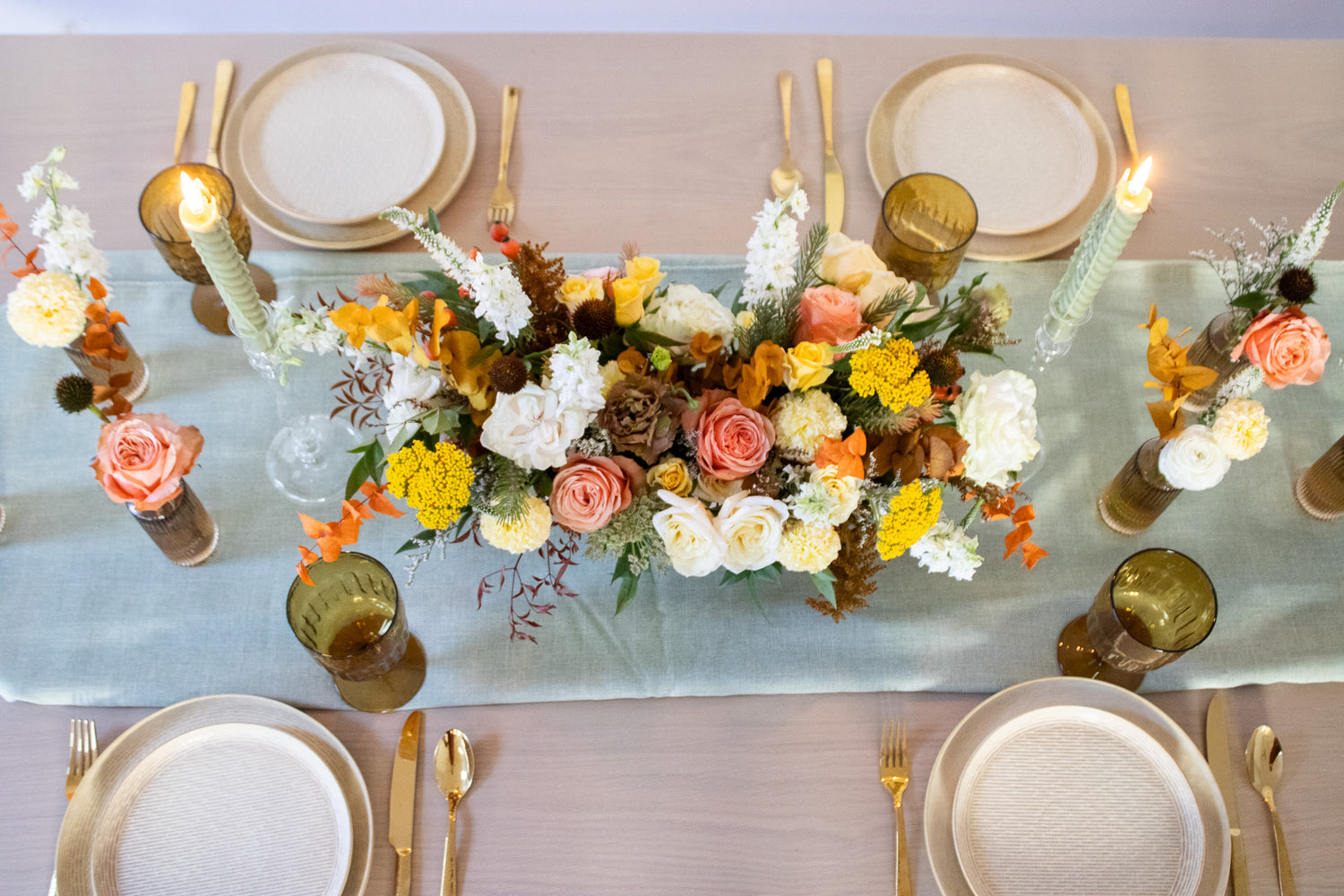 WildFlora's Gathered Garden Arrangement in an art deco white long & low container on a thanksgiving table. It include coral, yellow, gold, brown, orange, and white flowers and foliage, complete with roses, spray roses, weeping amaranthus, delphinium, lisianthus, echinacea, yarrow, and marigold. 