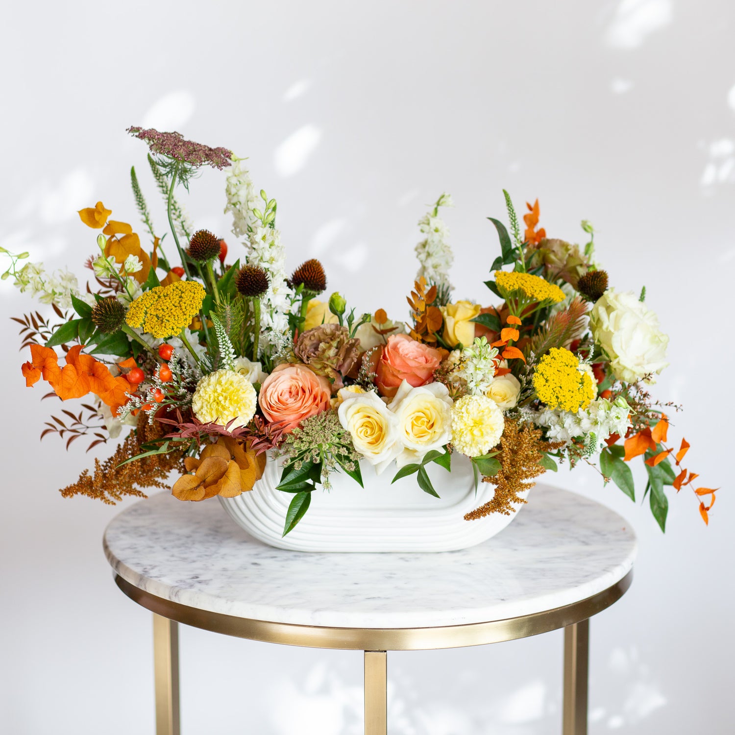 WildFlora's Gathered Garden Arrangement in an art deco white long & low container on a marble table. It include coral, yellow, gold, brown, orange, and white flowers and foliage, complete with roses, spray roses, weeping amaranthus, delphinium, lisianthus, echinacea, yarrow, and marigold. 