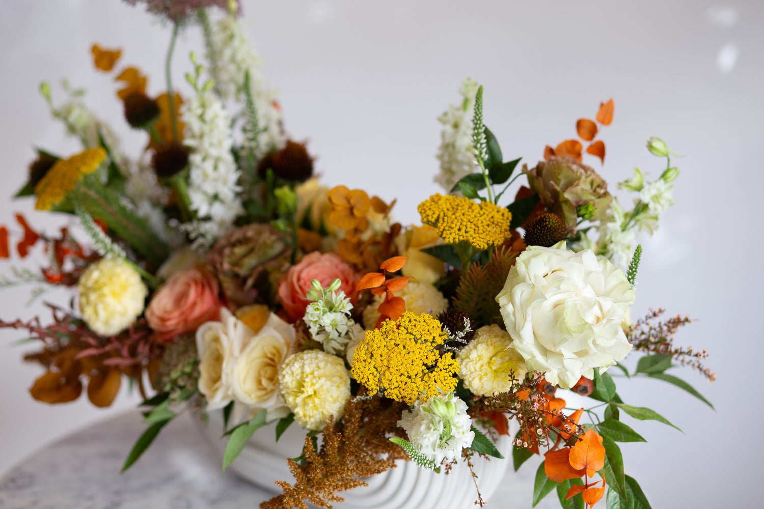 WildFlora's Gathered Garden Arrangement in an art deco white long & low container on a marble table. It include coral, yellow, gold, brown, orange, and white flowers and foliage, complete with roses, spray roses, weeping amaranthus, delphinium, lisianthus, echinacea, yarrow, and marigold. 