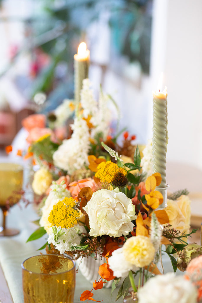 WildFlora's Gathered Garden Arrangement in an art deco white long & low container on a thanksgiving table. It include coral, yellow, gold, brown, orange, and white flowers and foliage, complete with roses, spray roses, weeping amaranthus, delphinium, lisianthus, echinacea, yarrow, and marigold. 