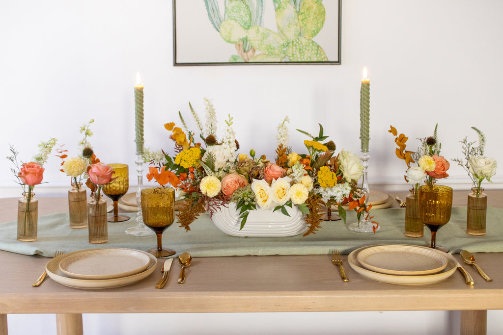 WildFlora's Gathered Garden Arrangement in an art deco white long & low container & Bud Vases in tan ribbed glass vases a on Thanksgiving table. They include coral, yellow, gold, brown, orange, and white flowers and foliage, complete with roses, spray roses, weeping amaranthus, delphinium, lisianthus, echinacea, yarrow, ranunculus, and marigold. 