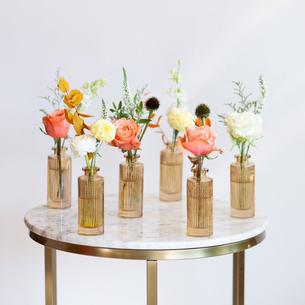 WildFlora's Gathered Garden Bud Vases in tan ribbed glass vases a on marble table. They include coral, yellow, gold, brown, orange, and white flowers and foliage, complete with roses, delphinium, veronica, echinacea, and ranunculus and marigold. 