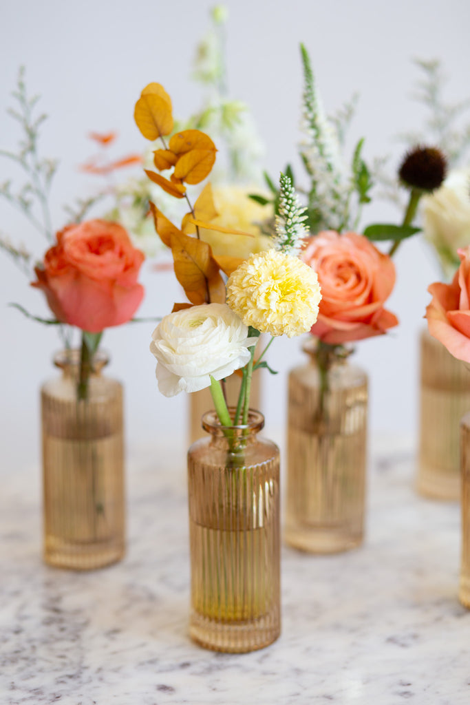 WildFlora's Gathered Garden Bud Vases in tan ribbed glass vases a on marble table. They include coral, yellow, gold, brown, orange, and white flowers and foliage, complete with roses, delphinium, veronica, echinacea, and ranunculus and marigold. 