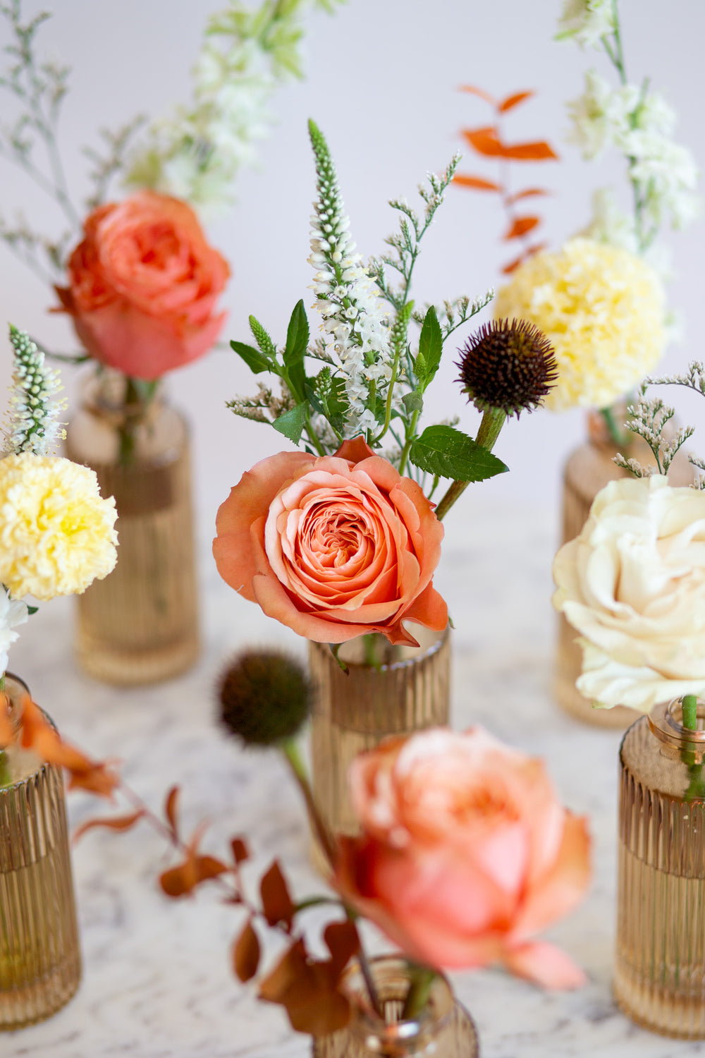 WildFlora's Gathered Garden Bud Vases in tan ribbed glass vases a on marble table. They include coral, yellow, gold, brown, orange, and white flowers and foliage, complete with roses, delphinium, veronica, echinacea, and ranunculus and marigold. 