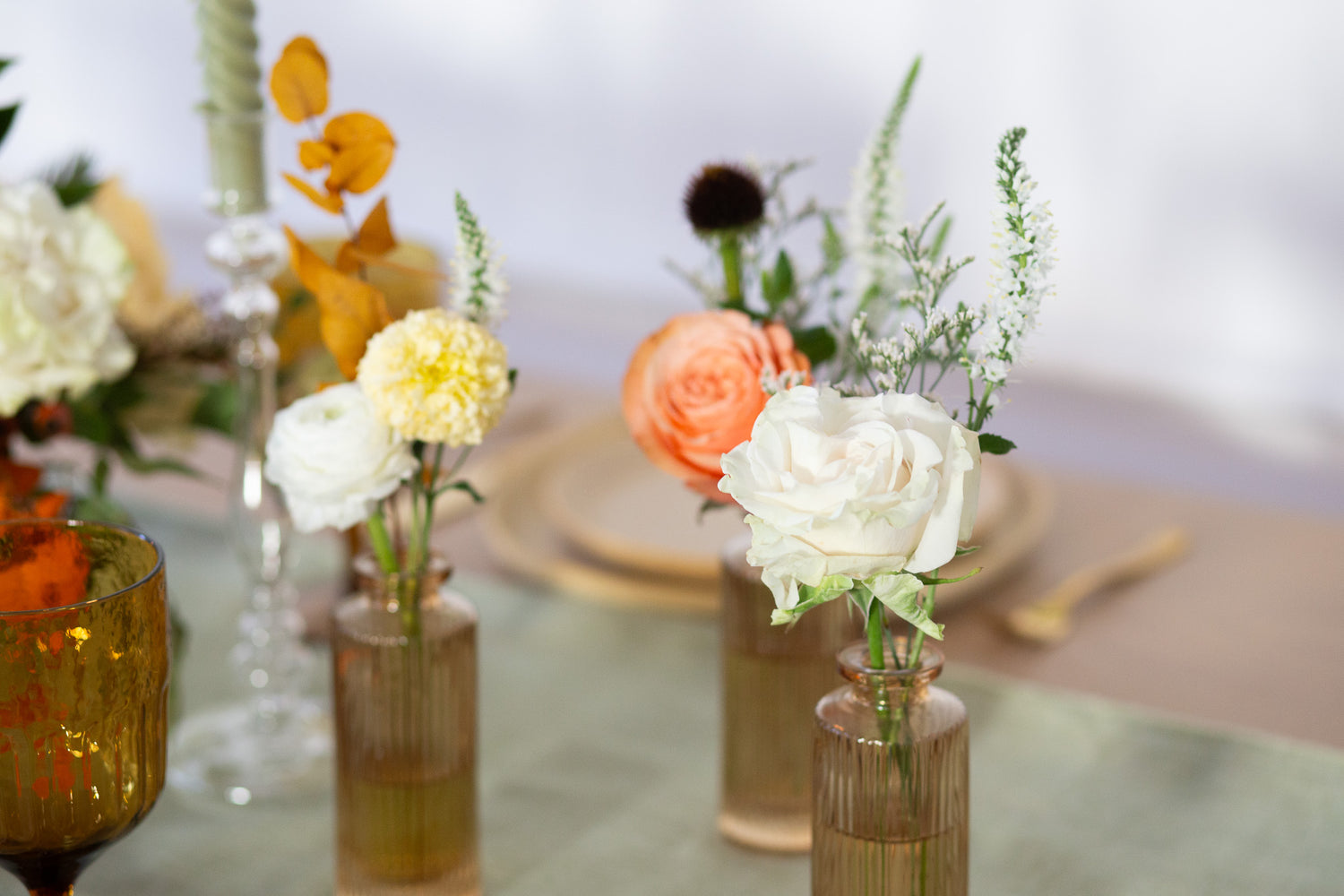 WildFlora's Gathered Garden Bud Vases in tan ribbed glass vases a on thanksgiving table. They include coral, yellow, gold, brown, orange, and white flowers and foliage, complete with roses, delphinium, veronica, echinacea, and ranunculus and marigold. 