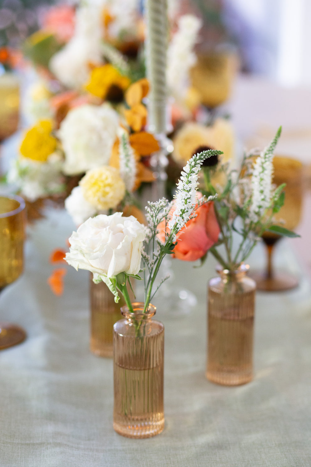 WildFlora's Gathered Garden Bud Vases in tan ribbed glass vases a on thanksgiving table. They include coral, yellow, gold, brown, orange, and white flowers and foliage, complete with roses, delphinium, veronica, echinacea, and ranunculus and marigold. 