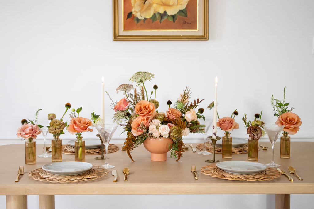 WildFlora's Gathered Garden Arrangement in a terracotta compote container & Bud Vases in tan ribbed glass vases a on Thanksgiving table. It includes dusty coral-orange, blush, brown, and tan earth-toned flowers, complete with Moab roses, spray roses, weeping amaranthus, Queen Anne's lace, lisianthus, echinacea, and foliage. 