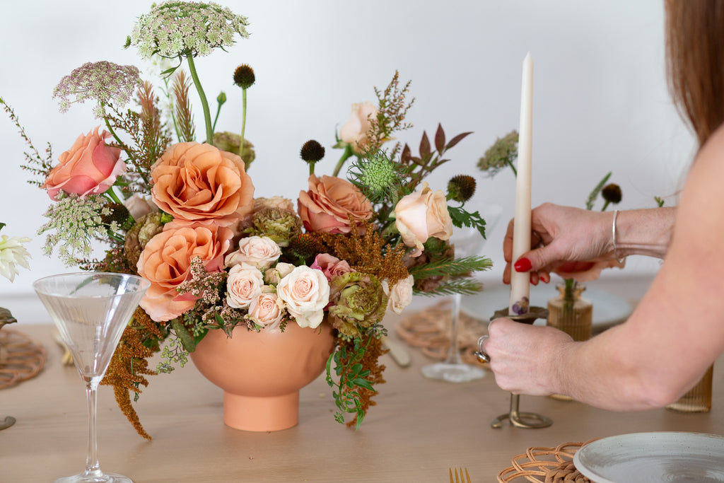 A woman sets a candle next to WildFlora's Gathered Garden Arrangement in a terracotta compote container & Bud Vases in tan ribbed glass vases a on Thanksgiving table. It includes dusty coral-orange, blush, brown, and tan earth-toned flowers, complete with Moab roses, spray roses, weeping amaranthus, Queen Anne's lace, lisianthus, echinacea, and foliage. 