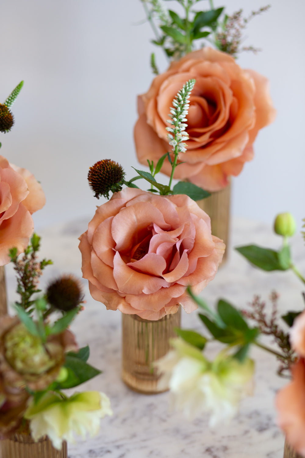 WildFlora's Gathered Garden Bud Vases in tan ribbed glass vases a on marble table. They include dusty coral-orange, blush, brown, and tan earth-toned flowers, complete with Moab roses, Queen Anne's lace, lisianthus, echinacea, and veronica.