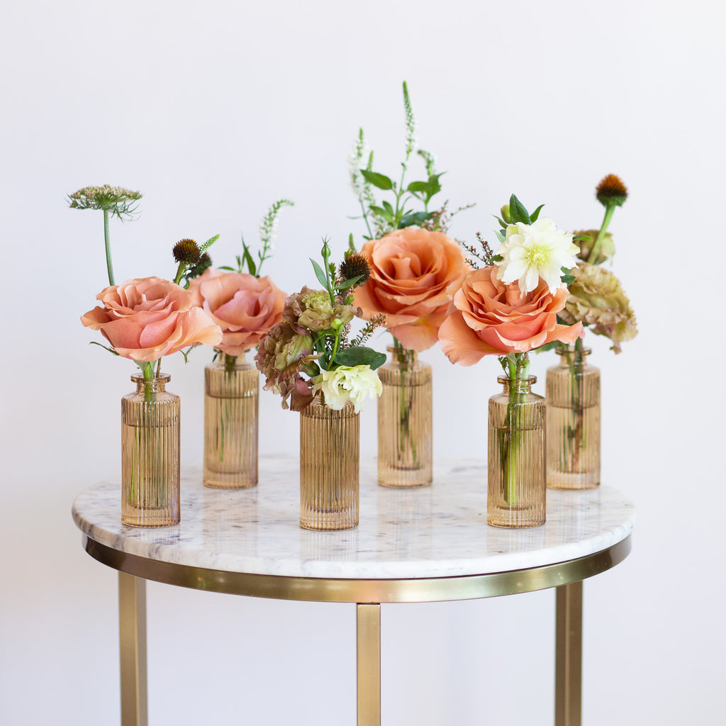 WildFlora's Gathered Garden Bud Vases in tan ribbed glass vases a on marble table. They include dusty coral-orange, blush, brown, and tan earth-toned flowers, complete with Moab roses, Queen Anne's lace, lisianthus, echinacea, and veronica.