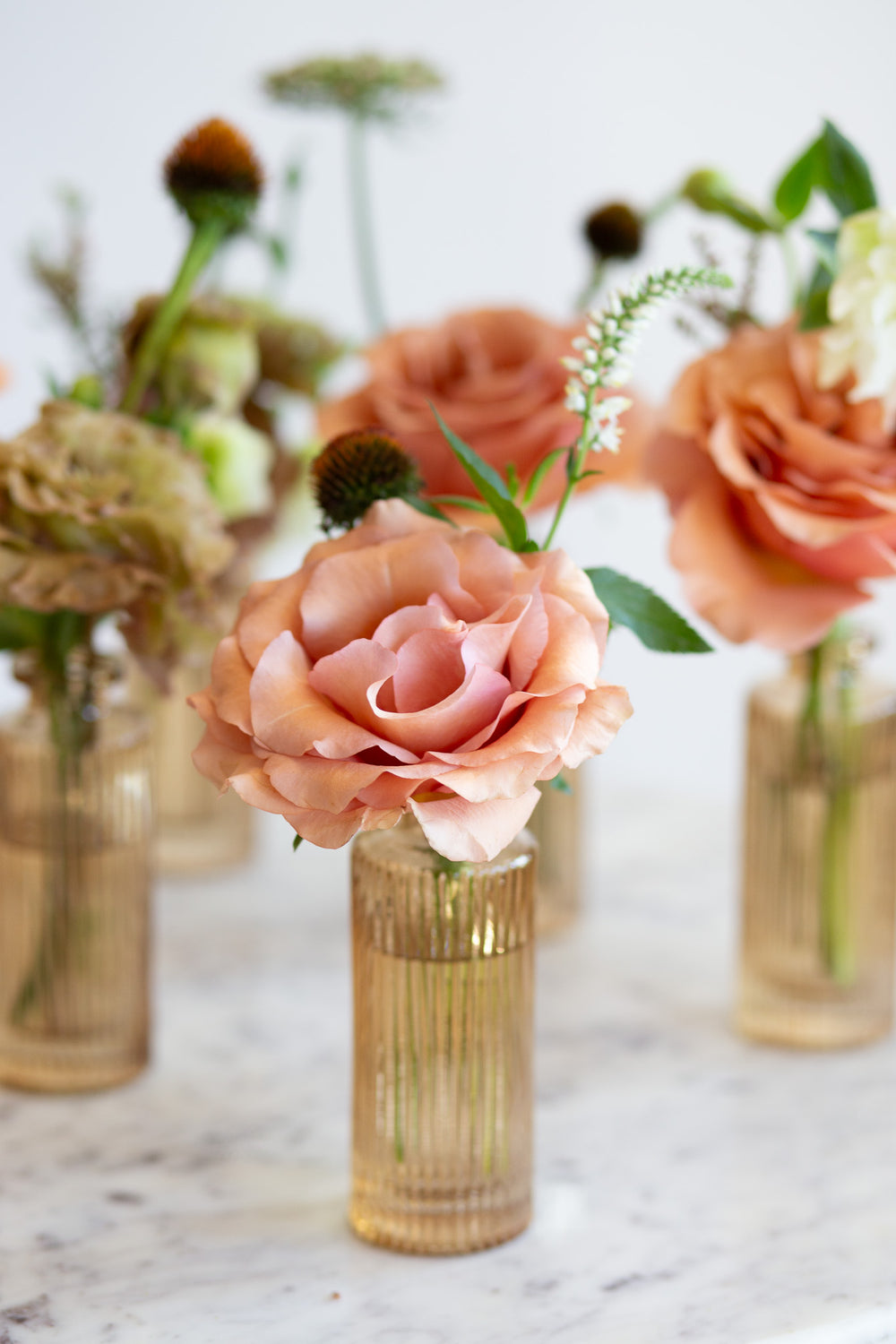 WildFlora's Gathered Garden Bud Vases in tan ribbed glass vases a on marble table. They include dusty coral-orange, blush, brown, and tan earth-toned flowers, complete with Moab roses, Queen Anne's lace, lisianthus, echinacea, and veronica.