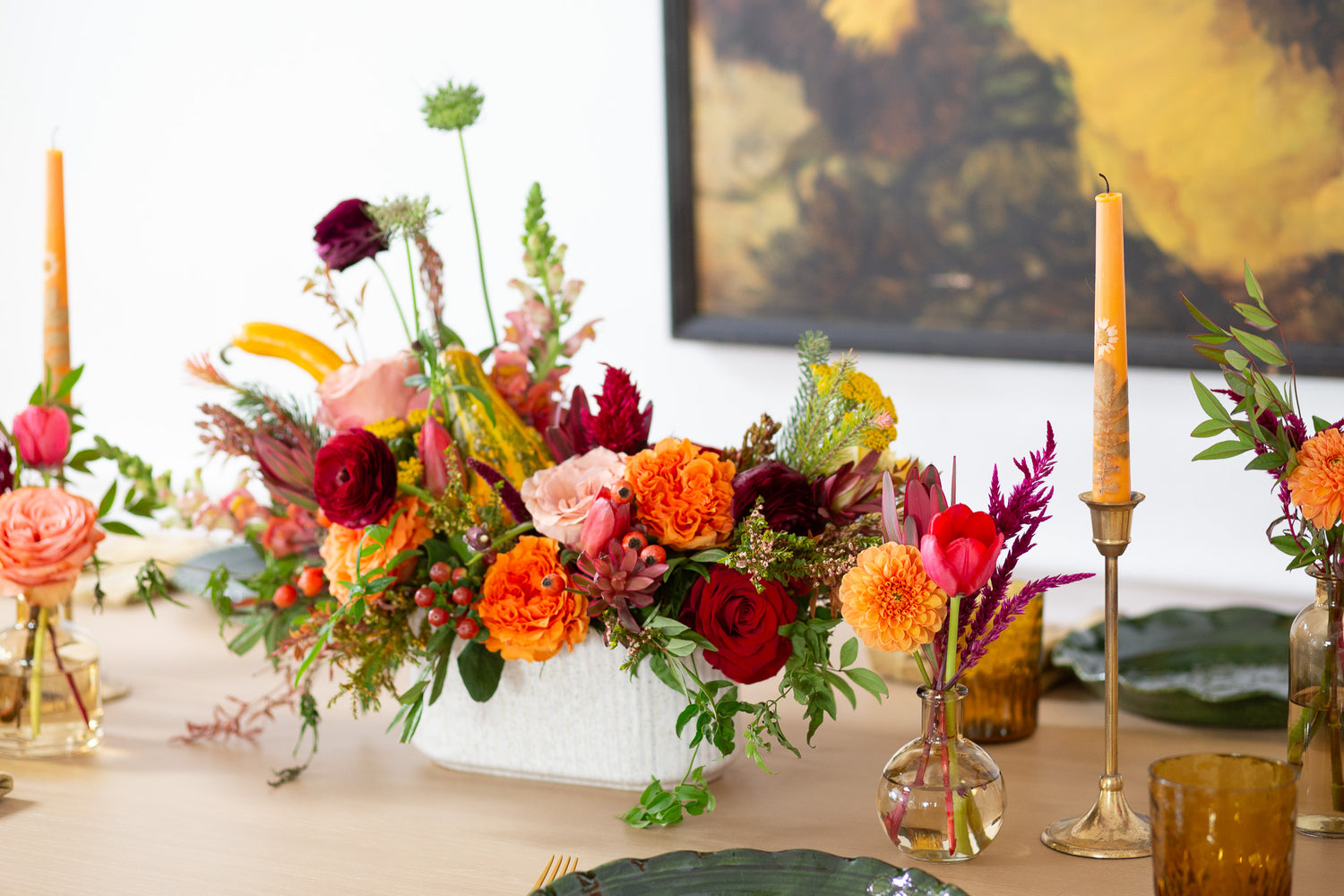 WildFlora's Gathered Garden Arrangement and Bud Vases in a ribbed long and low white speckled container on a Thanksgiving table. It includes orange, tangerine, red, pink, fuchsia, maroon, and gold flowers, complete with festive gourds / mini pumpkin, ranunculus, rose, dahlia, yarrow, weeping amaranthus, chocolate cosmos, delphinium, lisianthus, tulip, Queen Anne's lace, evergreens, snapdragon, and hypericum berries.