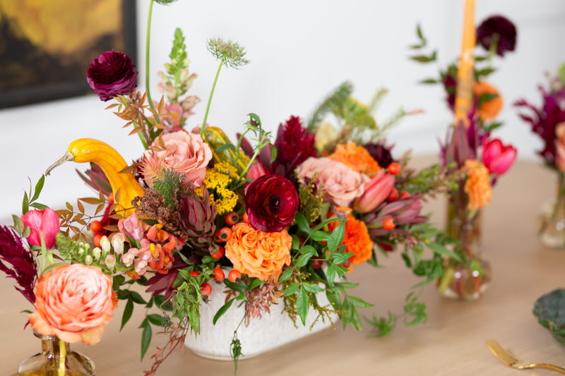 WildFlora's Gathered Garden Arrangement and Bud Vases in a ribbed long and low white speckled container on a Thanksgiving table. It includes orange, tangerine, red, pink, fuchsia, maroon, and gold flowers, complete with festive gourds / mini pumpkin, ranunculus, rose, dahlia, yarrow, weeping amaranthus, chocolate cosmos, delphinium, lisianthus, tulip, Queen Anne's lace, evergreens, snapdragon, and hypericum berries.